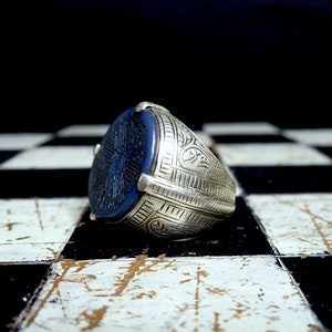 May include: A silver ring with a large, oval, dark blue stone. The stone is engraved with a detailed pattern. The ring's band is also decorated with intricate designs. The ring is displayed on a black and white checkered surface.