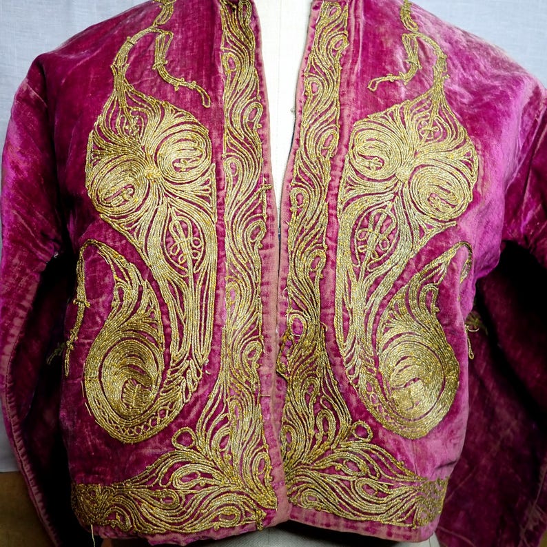 Pu&ograve; includere: Giacca in velluto magenta con ricami dorati. La giacca presenta motivi a spirale elaborati sul davanti e sui bordi, creando un design lussuoso e dettagliato. Le maniche sono visibili e la giacca &egrave; aperta sul davanti.