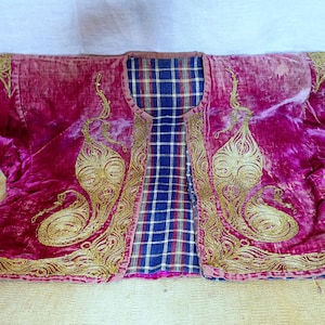 Pu&ograve; includere: Giacca in velluto magenta con ricami dorati. La giacca presenta una fodera a quadri blu, bianca e rossa. Il ricamo dorato presenta un motivo a paisley. La giacca &egrave; stesa su una superficie giallo chiaro.