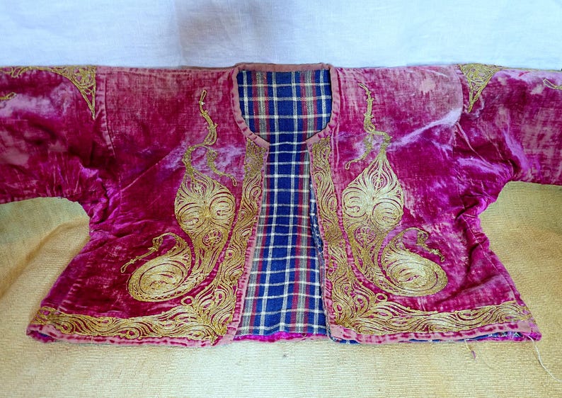 Pu&ograve; includere: Giacca in velluto magenta con ricami dorati. La giacca presenta una fodera a quadri blu, bianca e rossa. Il ricamo dorato presenta un motivo a paisley. La giacca &egrave; stesa su una superficie giallo chiaro.