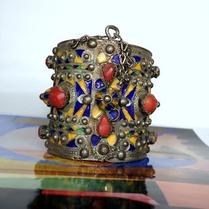 Può includere: Un bracciale a polsino in argento con un'intricata lavorazione a smalto in giallo, blu e verde. Il bracciale presenta accenti di pietre preziose rosse e un dettaglio a catena. Il design è ornato e dettagliato, in stile vintage o etnico.