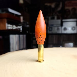 May include: A handcrafted, decorative object featuring a wooden, flame-shaped top with small, drilled holes. The top is a warm orange color and is attached to a brass base, creating a unique and artistic design.