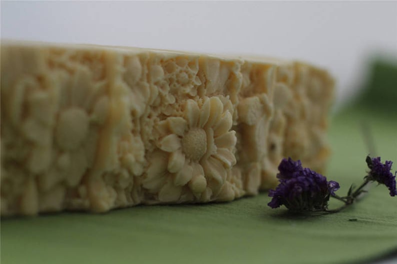 Pu&ograve; includere: Primo piano di una saponetta bianca con un design floreale. La saponetta &egrave; appoggiata su una superficie verde con fiori viola.