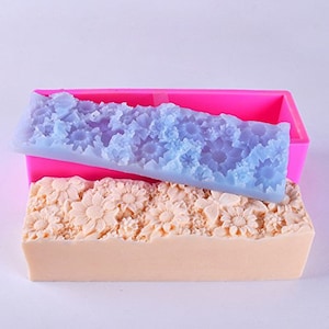 Op de afbeelding: Een roze siliconen mal met een bloemenpatroon. De mal is gevuld met een lichtblauwe zeepstaaf met een bloemenpatroon. De zeepstaaf ligt op een witte zeepstaaf met een bloemenpatroon.