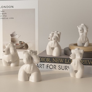 May include: Five white ceramic sculptures of female figures with abstract shapes and hearts on their chests. The sculptures are arranged on a table with a book and a wooden base.