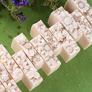 Pu&ograve; includere: Un set di otto saponette rettangolari con un design floreale. Il sapone &egrave; di colore beige chiaro e presenta un design in rilievo di fiori e foglie. Il sapone &egrave; disposto in fila su una superficie verde.