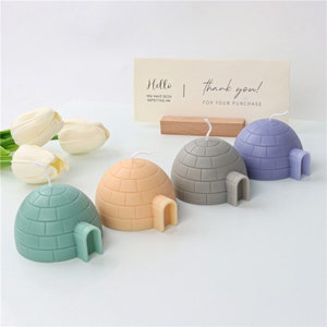May include: Four small igloo-shaped candles in different colors: green, beige, gray, and purple. The candles are arranged in a row on a white surface. A small wooden sign with the text "Hello, you have been expecting me" and "Thank you! For your purchase" is in the background.