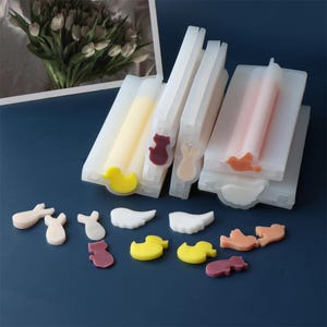 Puede incluir: Tres moldes de silicona para hacer jabón o velas. Los moldes son transparentes y tienen diferentes formas, incluyendo un pato, un gato, un conejo y un pájaro. Los moldes están sobre una superficie azul con varias piezas pequeñas de jabón o velas de colores.