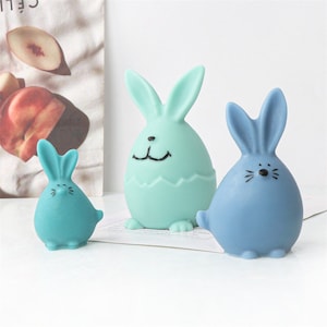 Könnte beinhalten: Drei Keramik-Osterhasen-Figuren. Der größte Hase ist hellgrün und hat eine weiße Eiform um sich herum. Der mittlere Hase ist hellblau und der kleinste Hase ist in einem dunkleren Blauton.