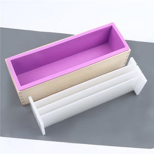 May include: A rectangular soap mold with a light wood exterior and a purple silicone interior. A white, multi-sectioned divider sits beside the mold. The mold is designed for making bar soap.