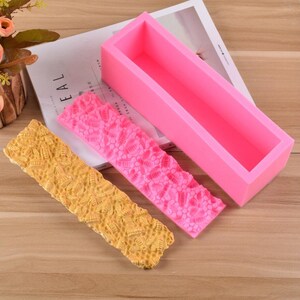May include: A pink rectangular silicone soap mold with two textured silicone mats. One mat is pink, the other is yellow. The mats have a repeating pattern of a floral design. The mold is empty.