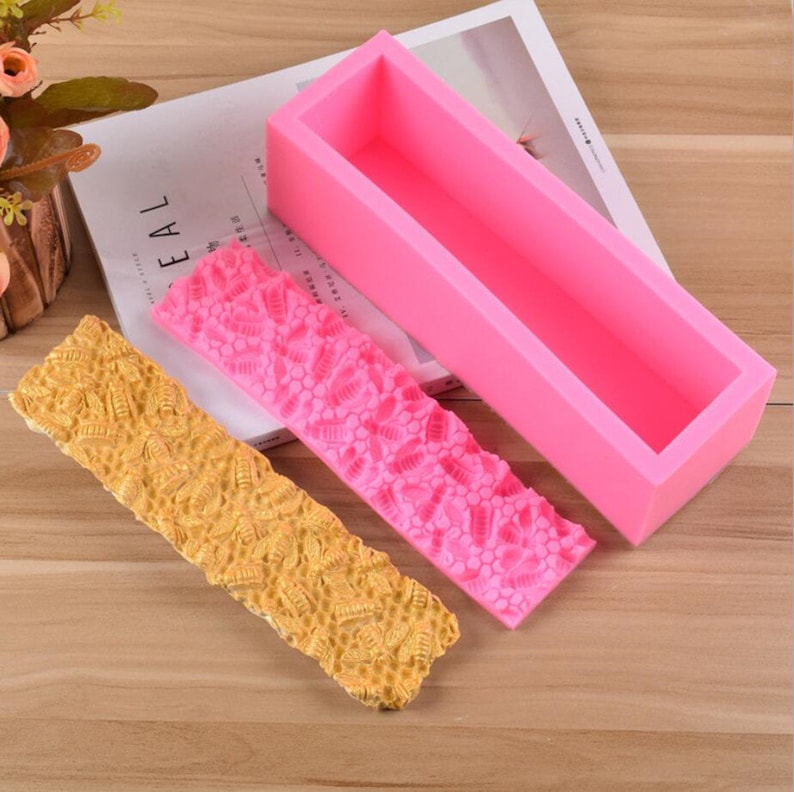 May include: A pink rectangular silicone soap mold with two textured silicone mats. One mat is pink, the other is yellow. The mats have a repeating pattern of a floral design. The mold is empty.