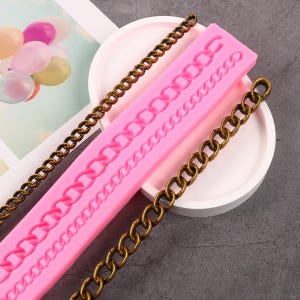 Puede incluir: Molde de silicona rosa con un diseño de cadena. El molde se utiliza para crear decoraciones de cadena comestibles para pasteles y otros postres. Una cadena dorada se muestra junto al molde.