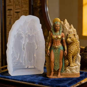 May include: A detailed statue of an Egyptian queen with a golden headdress, top, and armbands, and a teal skirt. A leopard sits beside her. A clear silicone mould of the queen and a dog is on the left. The statue is on a blue surface.