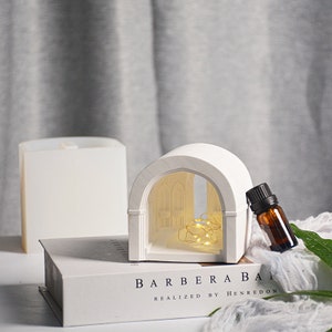 May include: White ceramic archway with a small window and a string of fairy lights inside. A small brown glass bottle with a dropper is next to the archway. The archway is sitting on a white book with the text "BARBERABAI REALIZED BY HENREDON".