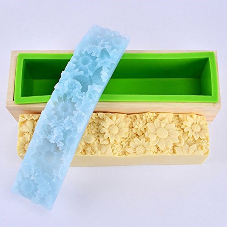 Pu&ograve; includere: Uno stampo per sapone in legno con un inserto in silicone blu chiaro e un inserto in silicone verde. Gli inserti in silicone hanno un motivo floreale. Una saponetta con un motivo floreale &egrave; appoggiata sullo stampo.