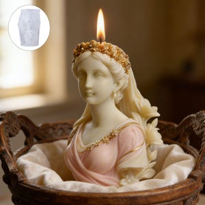Op de afbeelding: Een kaars in de vorm van een klassieke vrouwenbuste, met een brandende lont. De kaars is crèmekleurig met een roze en goudkleurige jurk en een bloemenkrans. Hij staat in een decoratieve houten mand, gevoerd met witte stof. Een doorzichtige mal staat linksboven.