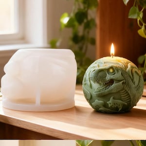 May include: A green dinosaur-shaped candle with a lit wick sits next to a white, dinosaur-shaped mold. The candle features detailed carvings of a T-Rex and other prehistoric elements. The candle is on a wooden surface.