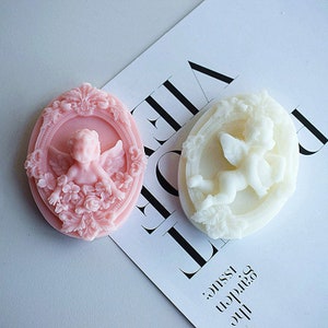 May include: Two oval-shaped decorative soap bars with a cherub design. The soap on the left is pink and the soap on the right is white.