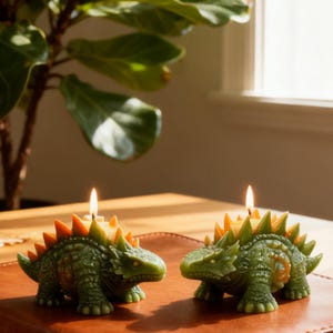 May include: Two dinosaur-shaped candles with lit wicks. The candles are green with orange and yellow accents, resembling a Stegosaurus. They are placed on a brown surface, with a plant in the background.