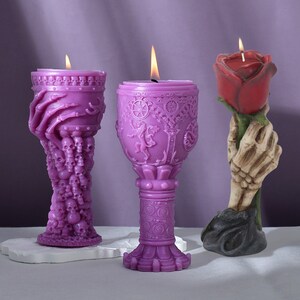 Puede incluir: Tres candelabros morados en forma de mano de calavera, copa y mano de esqueleto sosteniendo una rosa roja. Las velas están encendidas.