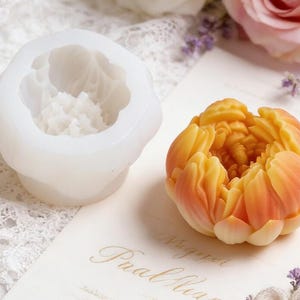 Puede incluir: Un molde de silicona blanco con forma de flor junto a una flor detallada, naranja y amarilla. El molde tiene un interior texturizado, y la flor está intrincadamente diseñada con capas de pétalos. El fondo incluye una rosa y delicadas flores moradas.