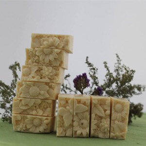 Pu&ograve; includere: Una pila di otto saponette rettangolari con un motivo floreale. Il sapone &egrave; di colore crema chiaro e presenta un delicato motivo floreale. Il sapone &egrave; disposto in pila su una superficie verde con fogliame verde sullo sfondo.