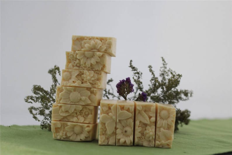 Pu&ograve; includere: Una pila di otto saponette rettangolari con un motivo floreale. Il sapone &egrave; di colore crema chiaro e presenta un delicato motivo floreale. Il sapone &egrave; disposto in pila su una superficie verde con fogliame verde sullo sfondo.