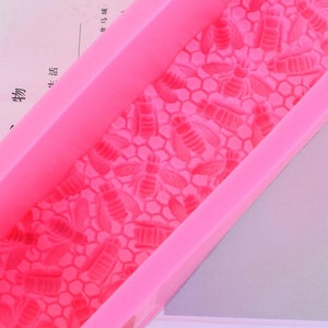 May include: A bright pink silicone mold featuring a honeycomb pattern with bee designs. The mold is rectangular with raised edges, perfect for crafting soaps, candles, or other DIY projects. The bee and honeycomb details are clearly defined.