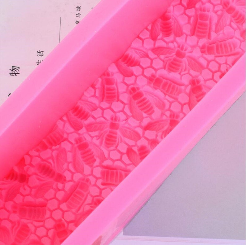 May include: A bright pink silicone mold featuring a honeycomb pattern with bee designs. The mold is rectangular with raised edges, perfect for crafting soaps, candles, or other DIY projects. The bee and honeycomb details are clearly defined.