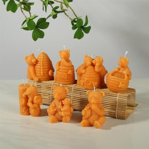 Puede incluir: Colección de velas con forma de oso naranja, algunas con panales o tarros de miel. Las velas están dispuestas sobre una esterilla de bambú, con follaje verde en el fondo. Las velas varían en tamaño y diseño, con mechas blancas.