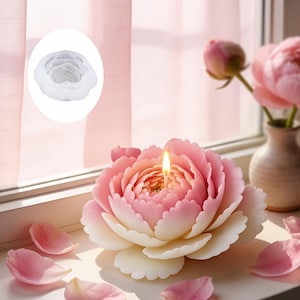 Puede incluir: Una vela con forma de peonía con una mecha encendida, con pétalos rosas y blancos. La vela está sobre un alféizar con pétalos esparcidos. Un pequeño jarrón con flores rosas está en el fondo.