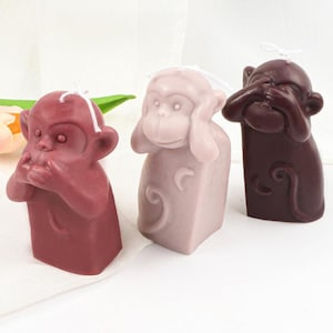 Könnte beinhalten: Drei Affen-Kerzenfiguren, jede mit einer anderen Handgeste. Der erste Affe bedeckt seinen Mund, der zweite seine Ohren und der dritte seine Augen. Die Kerzen sind aus braunem, rosa und violettem Wachs.