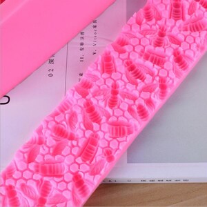 May include: A pink silicone mold featuring a honeycomb pattern with bee and leaf designs. The mold is designed for creating decorative elements for cakes, cookies, or other crafts. The mold is angled, showing the detailed texture.