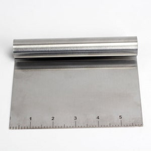 May include: A stainless steel pastry scraper with a ruler marked 1 to 5. The scraper has a straight edge and a rounded handle.