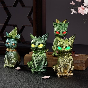May include: Four green and gold cat figurines with different designs. Each cat has glowing eyes and a unique design. The cats are sitting on a wooden surface.