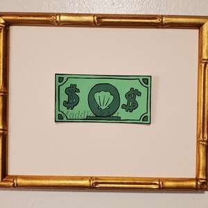 May include: A framed green paper bill with a seashell and dollar signs printed on it. The bill is titled "MaddJackArt".