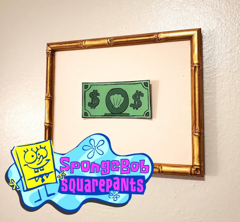 May include: A framed print of a green cartoon dollar bill with a seashell in the center. The frame is made of gold bamboo.