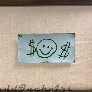 May include: A framed painting with a light blue background and a green smiley face with dollar signs on either side.