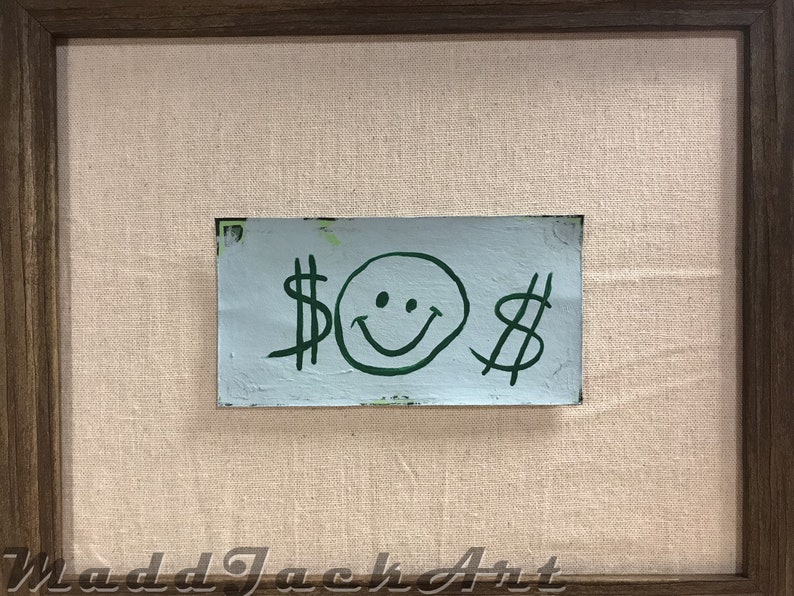 May include: A framed painting with a light blue background and a green smiley face with dollar signs on either side.