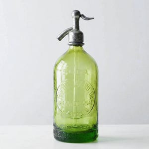 Pode incluir: Uma garrafa de sifão de soda de vidro verde vintage com um bico dispensador de metal prateado no topo. O corpo de vidro apresenta texto circular em relevo que diz SODA, PUERTO LIZANDRIA, UDAONDO 4541, LANUS e INDUSTRIA ARGENTINA.