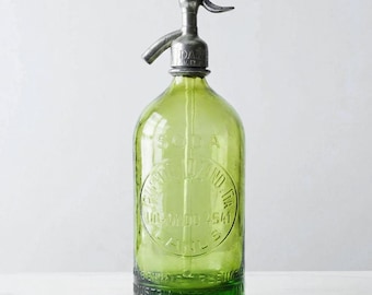 May include: A vintage green glass soda siphon bottle featuring a metallic silver-toned dispensing nozzle at the top. The glass body is embossed with circular text that reads SODA, PUERTO LIZANDRIA, UDAONDO 4541, LANUS, and INDUSTRIA ARGENTINA.