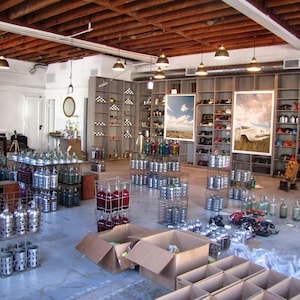 May include: A large warehouse space filled with numerous metal wire crates containing glass bottles in various colours, including blue, red, and clear. Tall wooden shelving units line the back wall, displaying assorted items and two large framed prints.