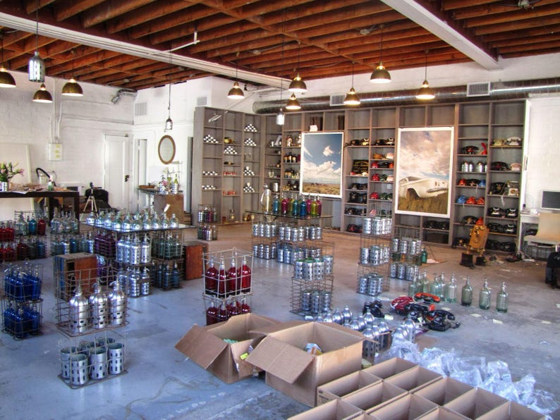 May include: A large warehouse space filled with numerous metal wire crates containing glass bottles in various colours, including blue, red, and clear. Tall wooden shelving units line the back wall, displaying assorted items and two large framed prints.