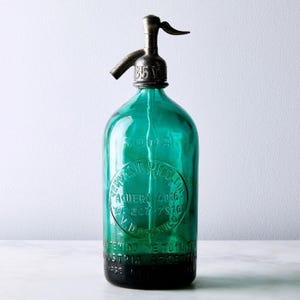 Pode incluir: Uma garrafa de sifão de soda vintage de vidro verde esmeralda com um bico de metal prateado. O vidro apresenta texto em relevo, incluindo SODA, GERVASIO RICO, AGUERO 4016, TE 207-7910 e INDUSTRIA ARGENTINA. O mecanismo superior é uma alavanca.