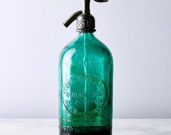 Pode incluir: Uma garrafa de sifão de soda vintage de vidro verde esmeralda com um bico de metal prateado. O vidro apresenta texto em relevo, incluindo SODA, GERVASIO RICO, AGUERO 4016, TE 207-7910 e INDUSTRIA ARGENTINA. O mecanismo superior é uma alavanca.