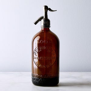 Pode incluir: Uma garrafa de sifão de soda de vidro âmbar vintage com um bico e mecanismo de alavanca de metal prateado. O vidro apresenta texto em relevo que diz SODA, P.T. LAVALLEN, 500 DE ESTERO 4046, LANUS, INDUSTRIA ARGENTINA.