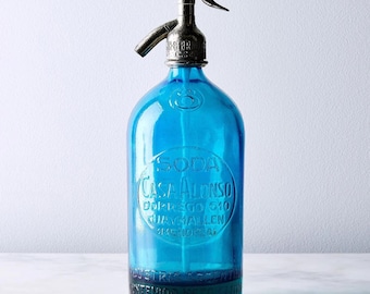 Pode incluir: Uma garrafa de sifão de soda de vidro azul brilhante vintage com um bico dispensador de tom prateado metálico. O corpo de vidro apresenta o texto em relevo SODA CASA ALONSO DORREGO 510 GUAYMALLEN MENDOZA e INDUSTRIA ARGENTINA.