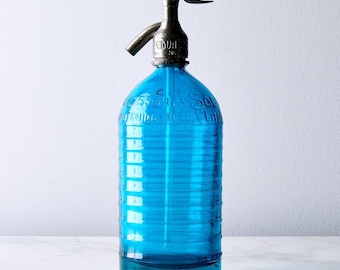 May include: A vintage blue glass soda siphon bottle with a metallic silver-toned nozzle and handle. The cylindrical glass body features horizontal ribbed detailing and embossed text that includes the phrase CONTENIDO NETO 1 LITRO.