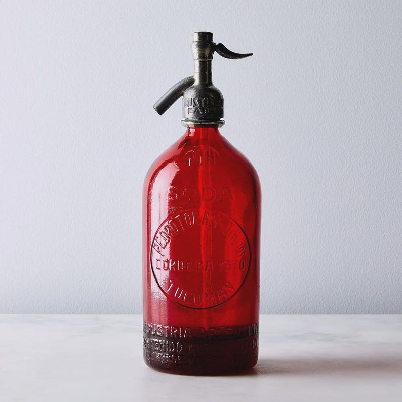 May include: A vintage red glass soda siphon bottle featuring a metallic dispensing nozzle at the top. The glass body is embossed with circular text reading Pedro Tomas, Cordoba 1370, Tucuman, and Industria Argentina at the base.